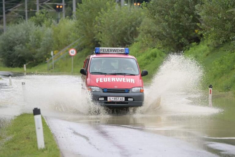 Einsatzfahrzeug Last fährt durch tiefe Pfütze während Hochwassereinsatz in Fritzens. Wasserfontänen spritzen seitlich hoch.